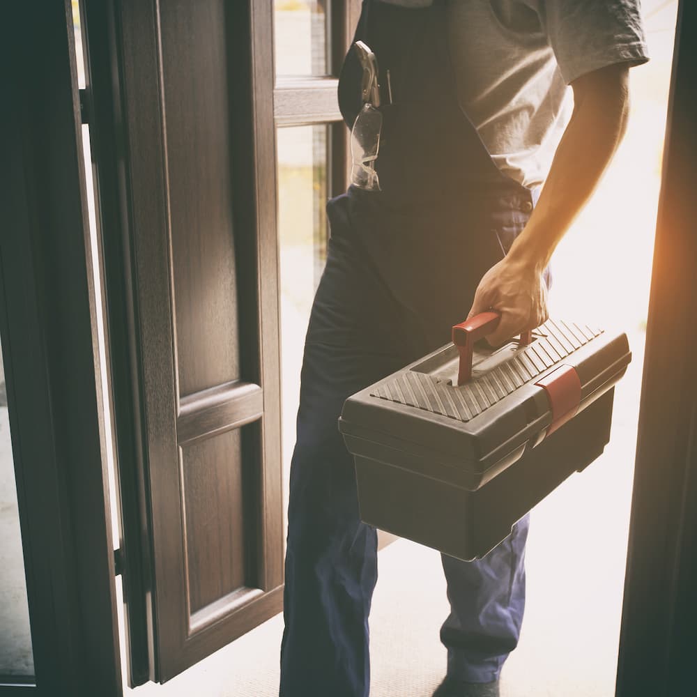 A man wearing cover-alls and holding a tool-box enters through the front door of the home.